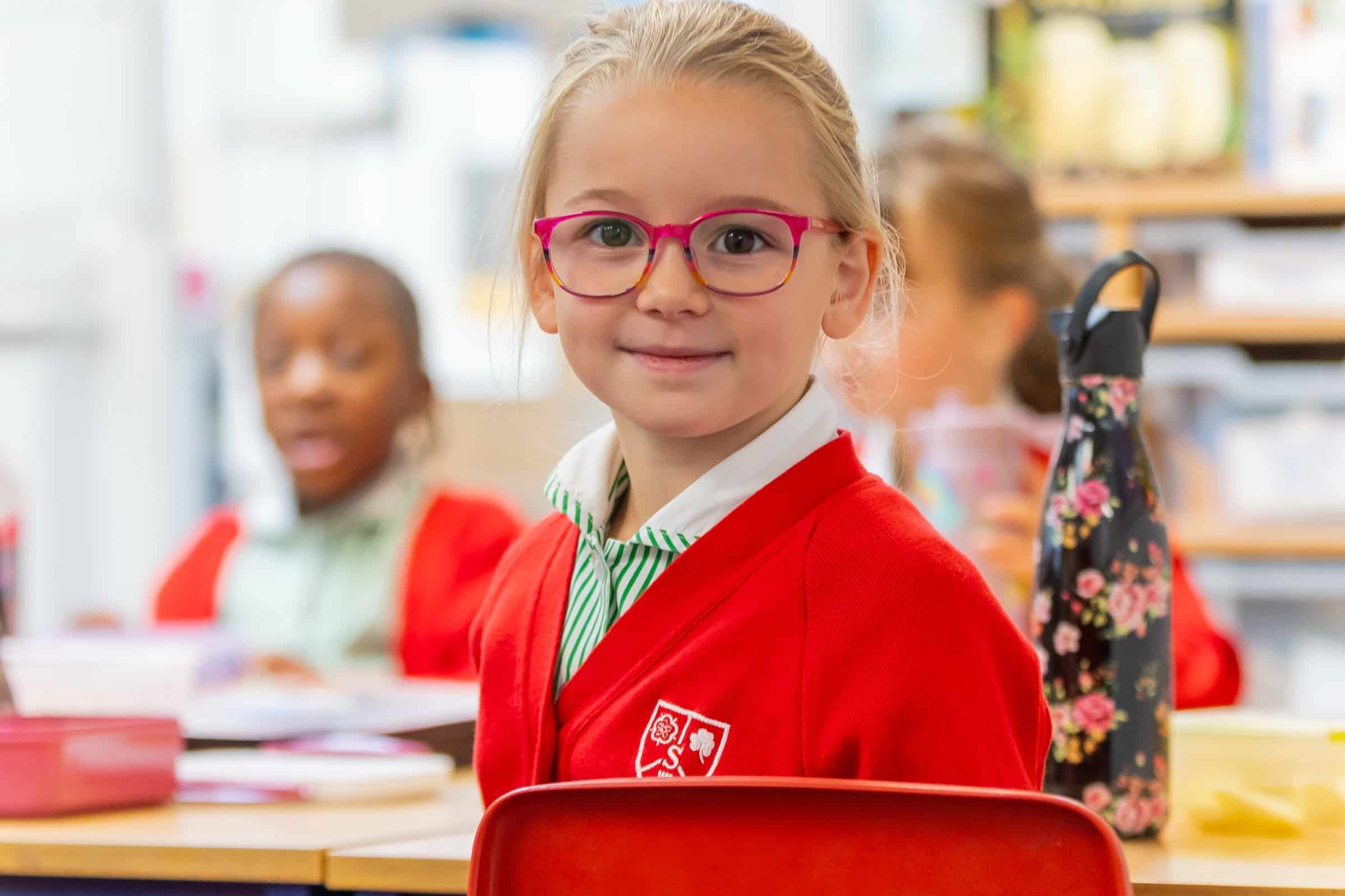 Young girl with blonde hair and pink glasses, wearing a red school uniform, smiles whilst sitting at her desk in a classroom—a proud future member of the Stormont Old Girls. Other children and classroom materials are visible in the background.