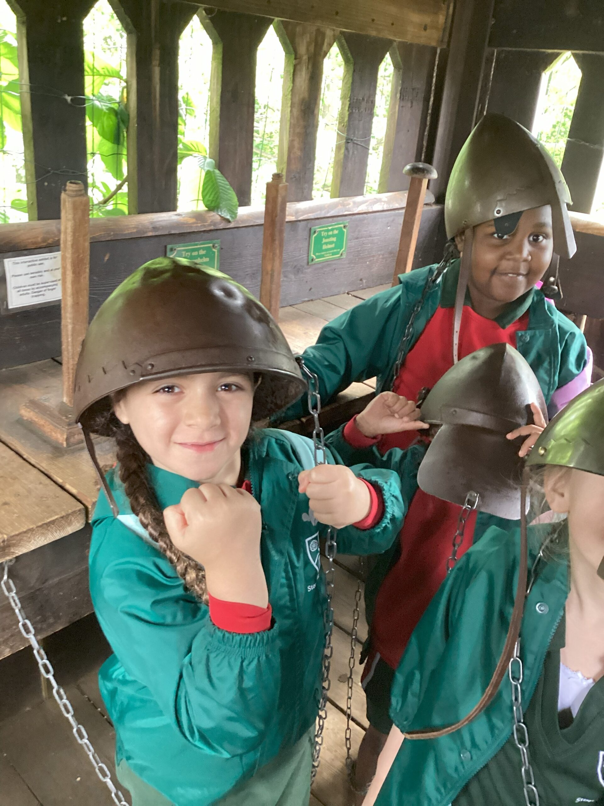 Children wearing medieval-style helmets and green jackets smile and pose with their fists up inside a wooden structure, taking part in a historical or educational activity.
