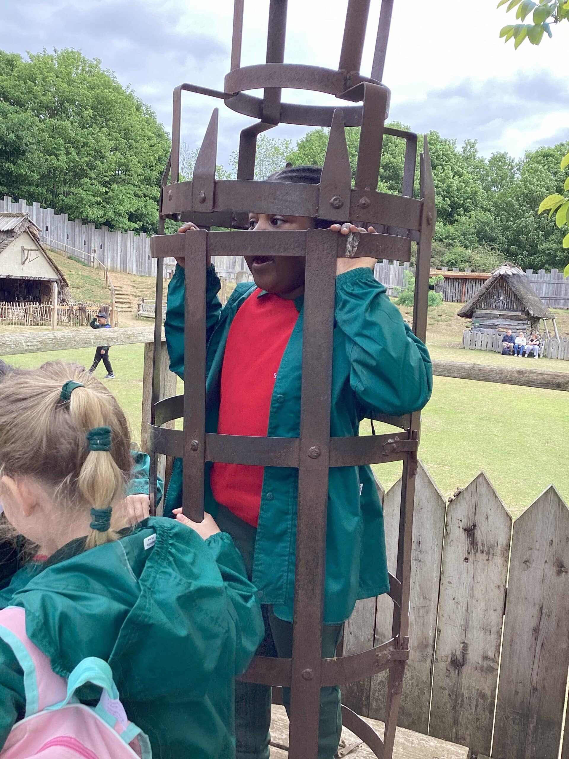 A child in a red shirt and green jacket stands inside a metal cage structure outdoors, with another child nearby. There are wooden fences, grassy areas, and historic-style huts in the background.
