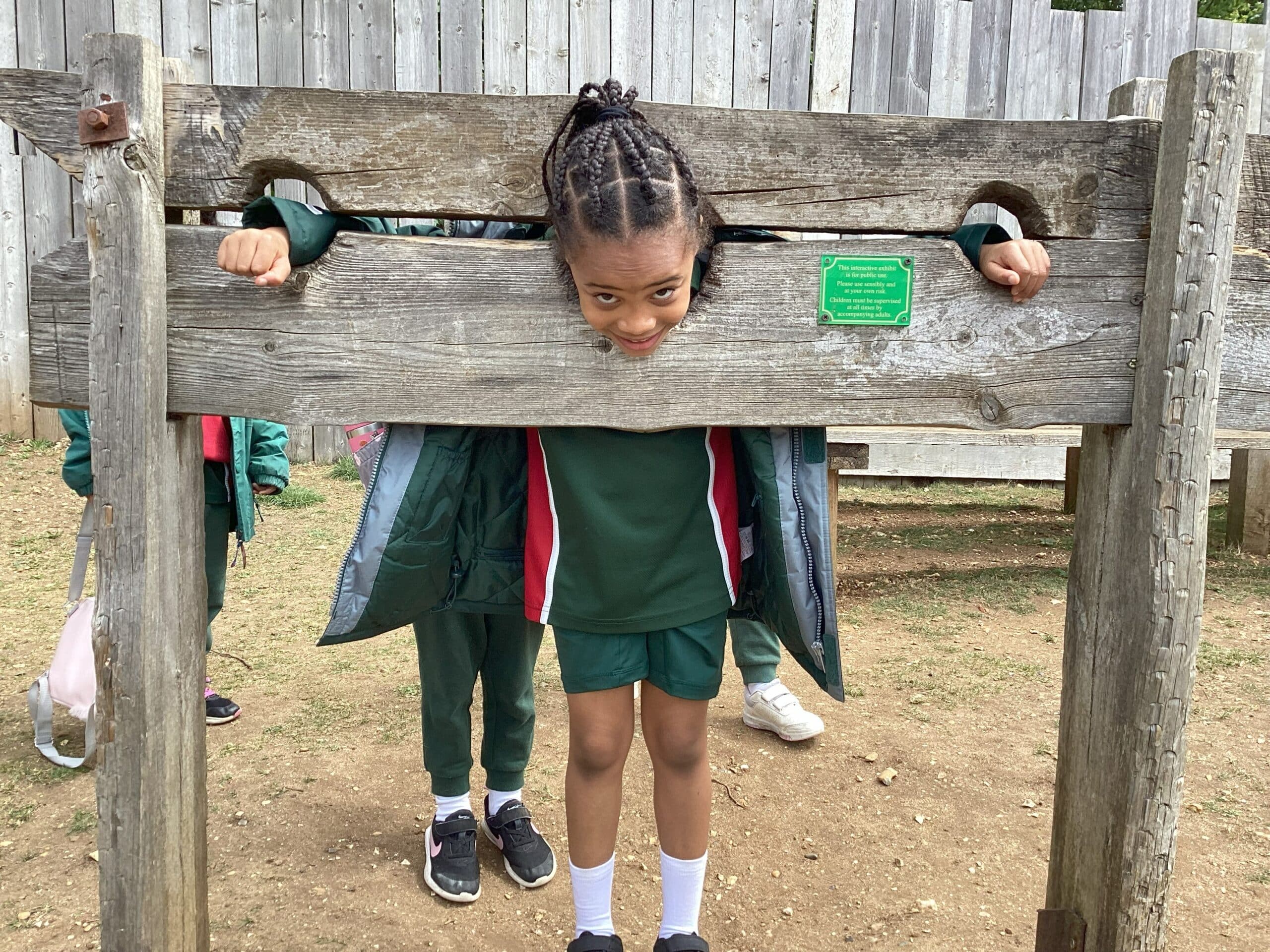 A young girl in a green school uniform stands with her head and hands in wooden stocks at an outdoor historical site, smiling playfully at the camera. Other children and a wooden fence are in the background.