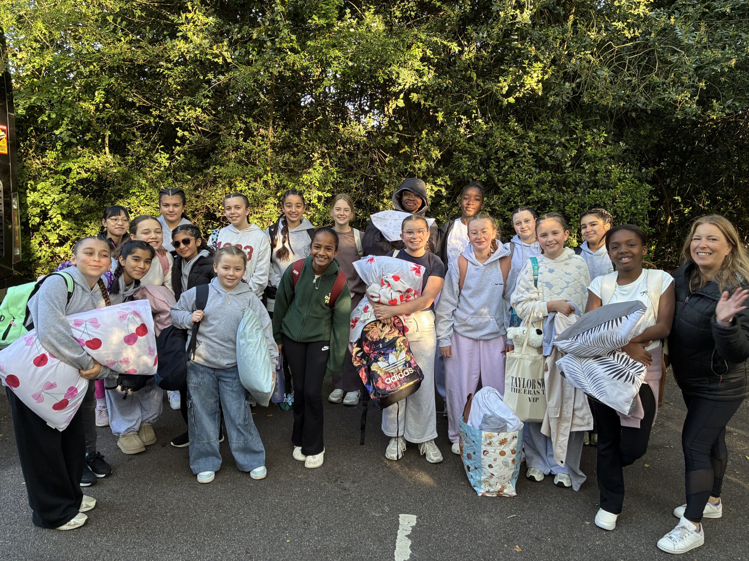A group of smiling young people and one adult stand outdoors holding pillows and bags, dressed in casual or pyjama-like clothes, with trees in the background. They appear happy and ready for an outing or sleepover.