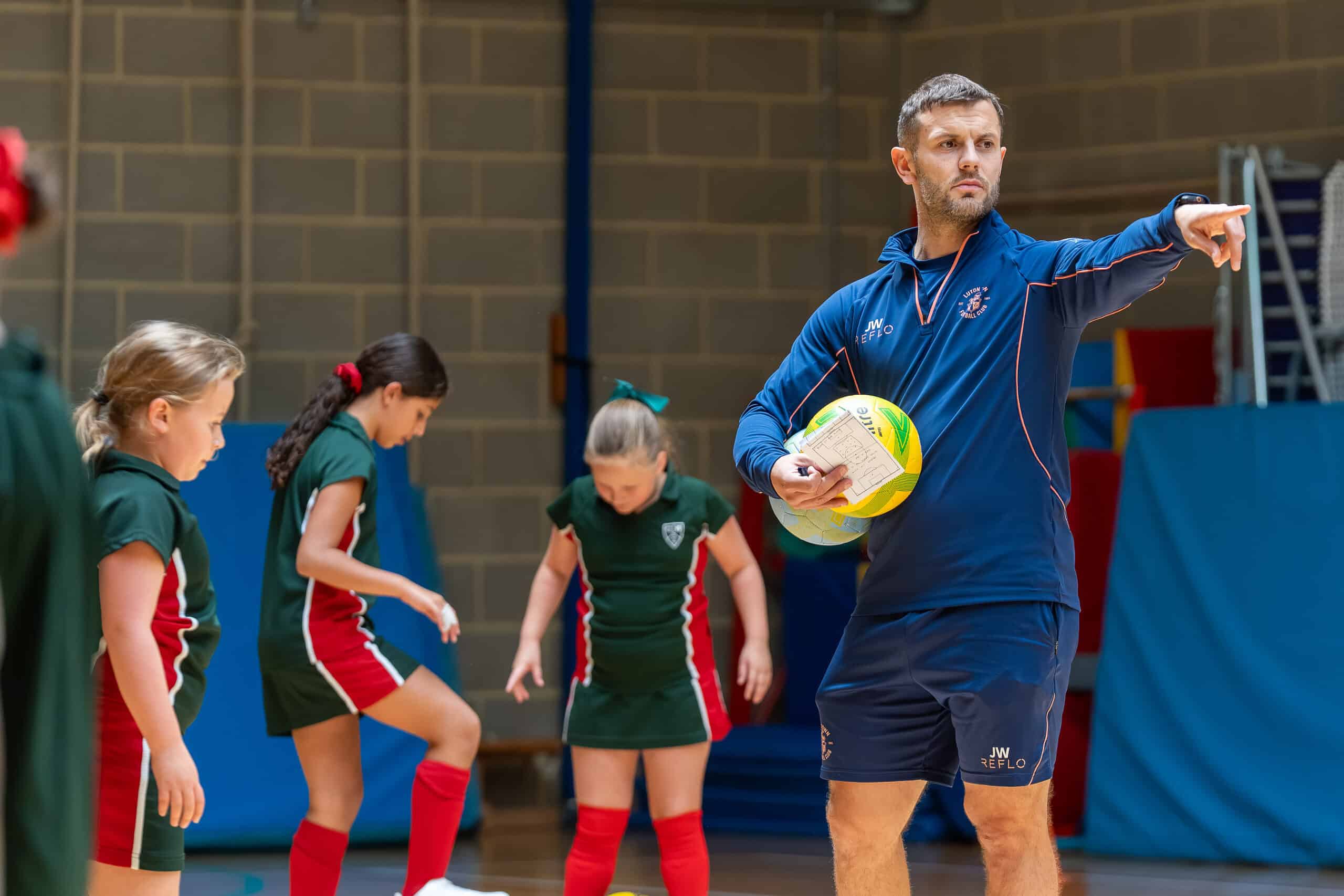 A male coach holding a ball gives instructions to young girls in green sports kits during an indoor practice session. Some girls are standing and listening, while others look down, possibly preparing for an activity.
