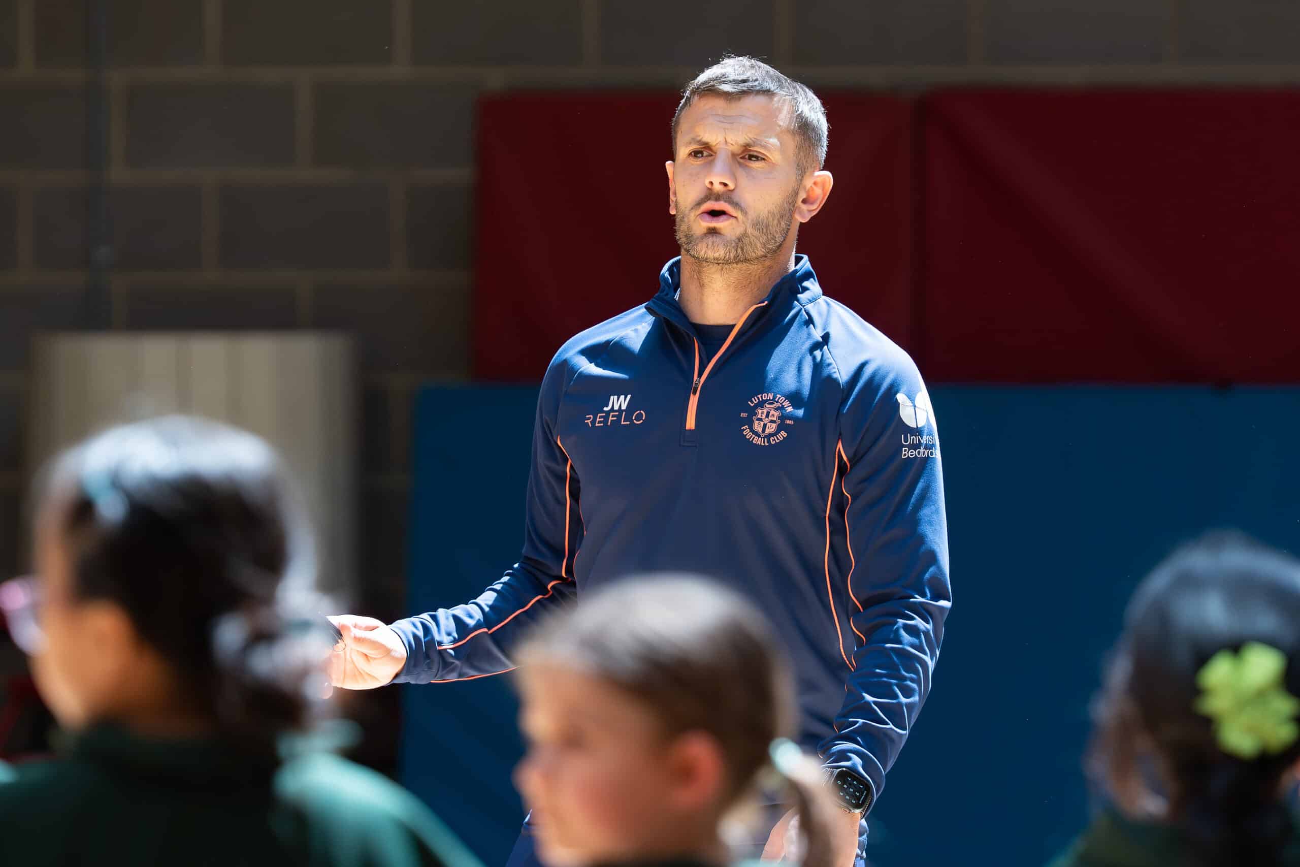 A man in a blue tracksuit top speaks animatedly, gesturing with his hands, while children in green uniforms listen in an indoor setting with gym mats on the wall behind him.