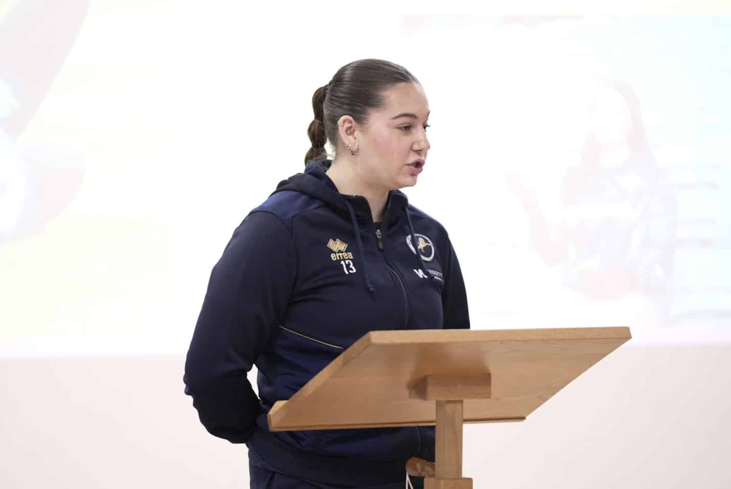 A woman in a navy sports tracksuit stands behind a wooden lectern, speaking. The background is plain with a faint, blurred image projected on the wall.