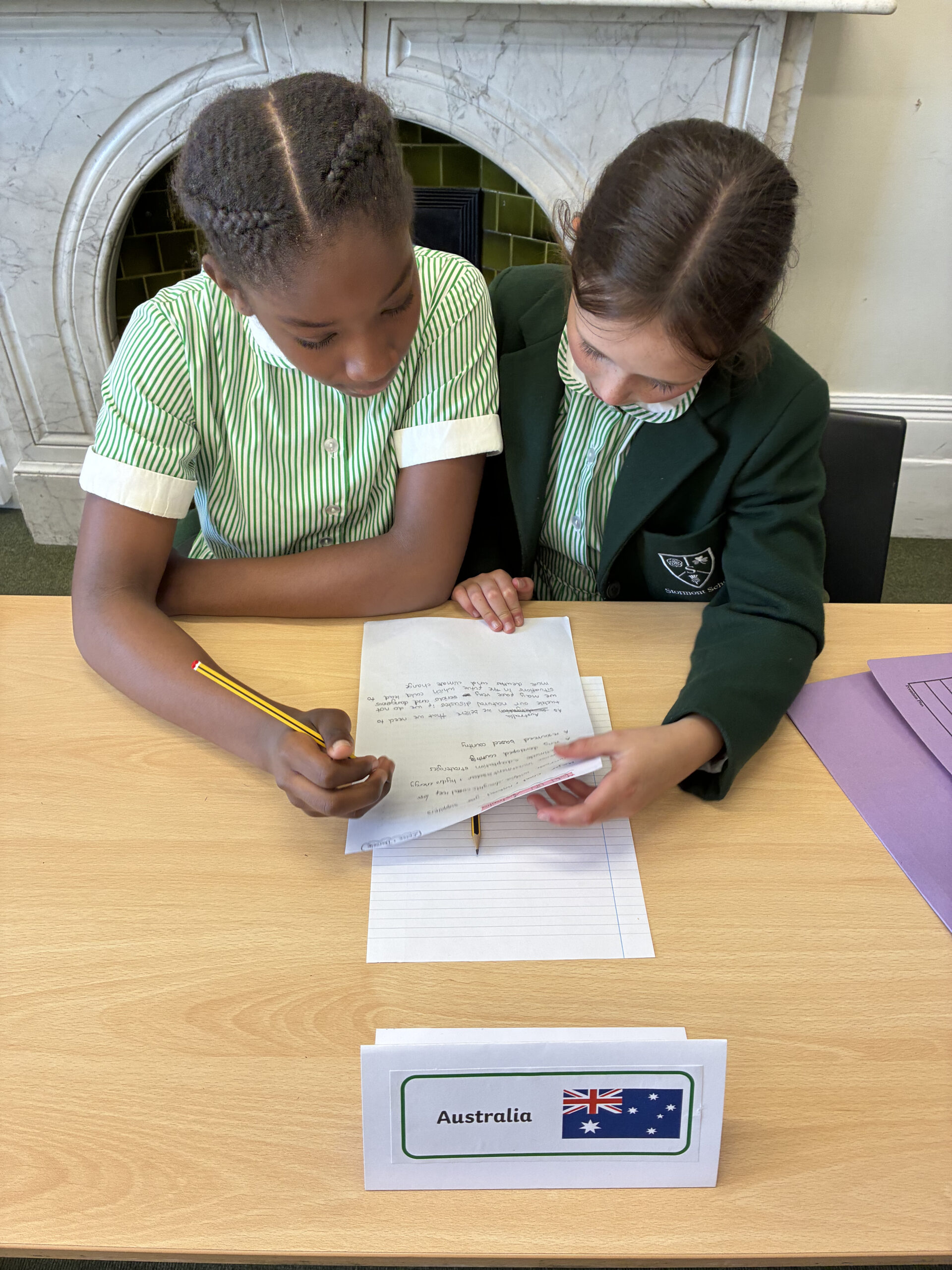 Two schoolgirls in green uniforms sit at a desk, working together on a piece of paper. In front of them is a sign labelled Australia with the Australian flag.