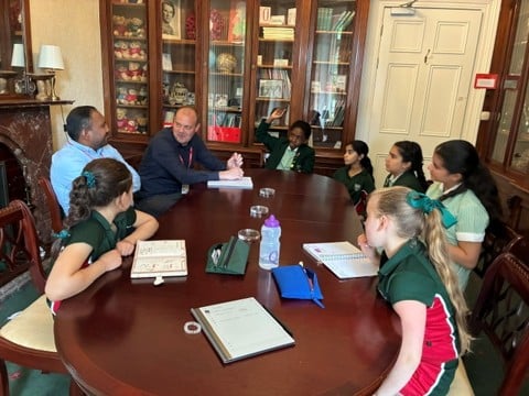A group of students in green uniforms and two adults sit around a wooden table in a room with bookshelves, discussing and taking notes. One student has her hand raised.