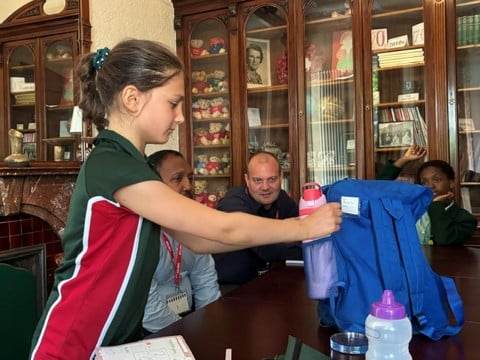 A young girl places a water bottle into a blue rucksack on a table whilst three people watch her in a room with glass cupboards filled with books and collectables.