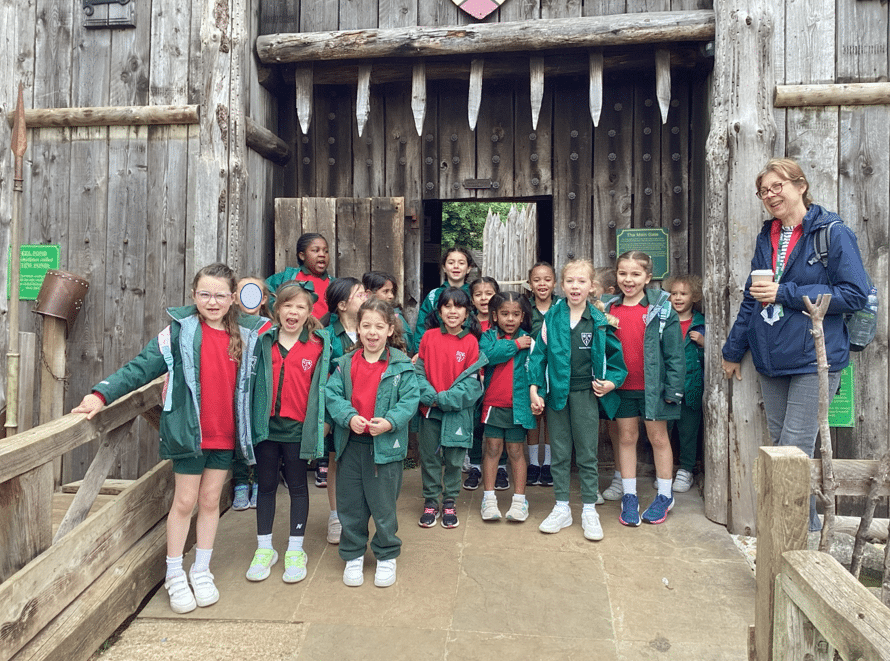 A group of smiling schoolchildren in green uniforms and red shirts stand in front of a wooden fort entrance, accompanied by an adult holding a coffee cup.