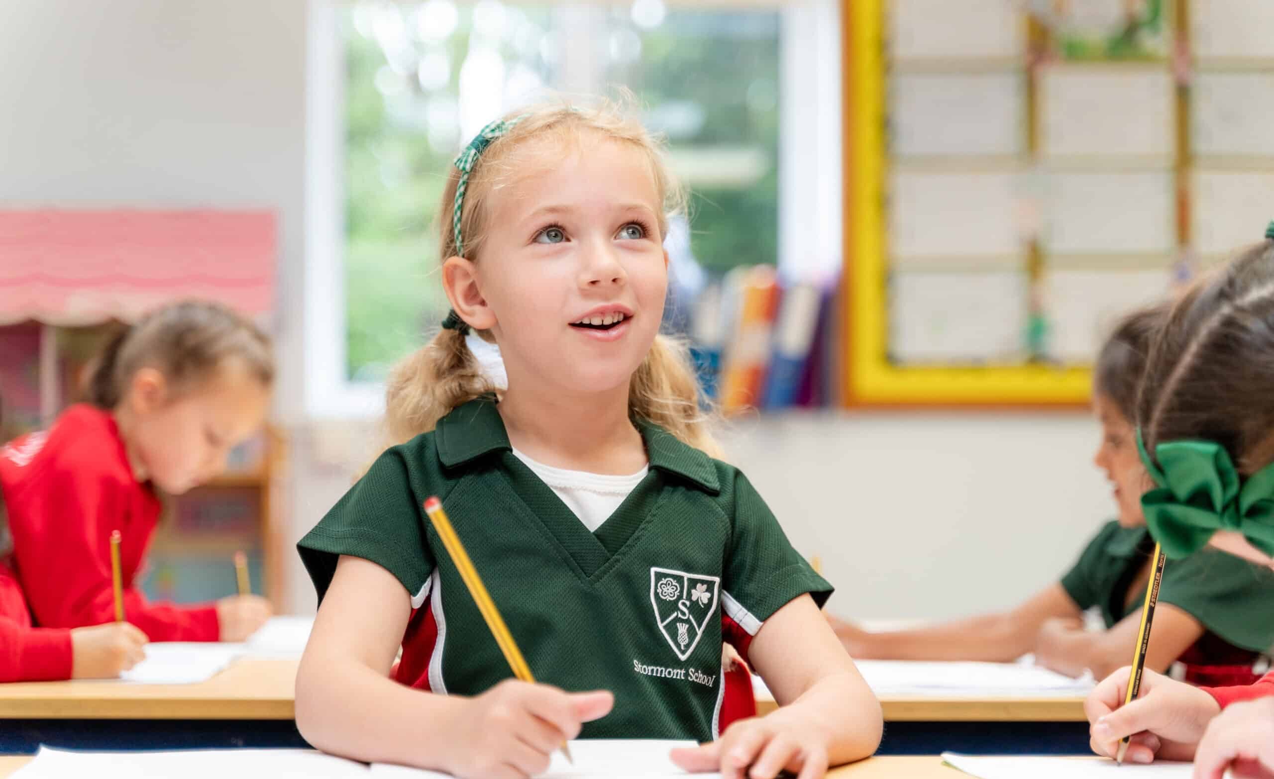 A young girl with blonde hair in plaits, wearing a green Stormont School uniform, smiles whilst sitting at a classroom desk with a pencil and exercise book. Other children are also working at their desks in the background.