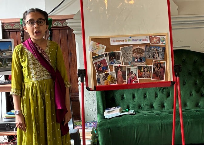 A young girl in a green traditional outfit stands beside an easel displaying a collage of photographs and a sign reading “A Journey to the Heart of India.” She appears to be giving a presentation in a sitting room, reminiscent of the festive spirit at Potters Bar Carnival.