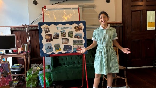A young girl in a green striped dress stands smiling next to a display board with photos and the word Thailand during a presentation at the Potters Bar Carnival, in a room with wooden walls and chairs.