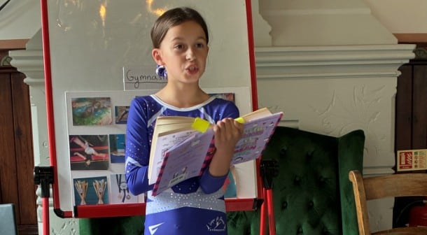 A young girl in a blue gymnastics outfit stands in front of a whiteboard, holding an open book and speaking. The whiteboard features photos and the word Gymnastics—she looks to be giving a presentation, perhaps preparing for the Potters Bar Carnival.