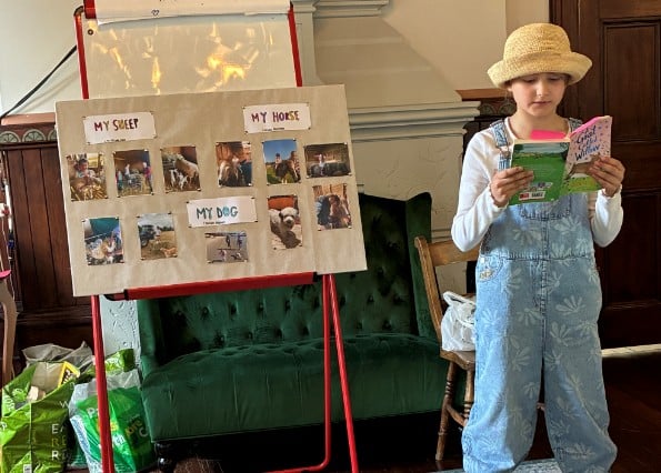 A young girl in dungarees and a straw hat reads from a book beside a display board labelled MY SHEEP, MY HORSE, and MY DOG at the Potters Bar Carnival. A green sofa and shopping bags are visible in the background.